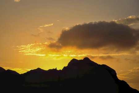 Silhouette of mountain ridge at sunset with golden sky and dramatic clouds.の写真素材