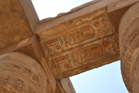 Close-up of a stone beam with colorful ancient Egyptian hieroglyphs inside a temple, supported by decorated sandstone columns under a clear blue sky.の写真素材