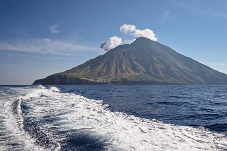 View of the Stromboli volcano with smoke rising from the crater, seen from the sea with boat wake in the foreground under a clear blue sky.の写真素材