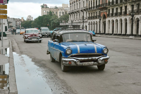 Vintage American cars driving through a historic street in Havana, Cuba. Old buildings line the road on a cloudy day.の写真素材
