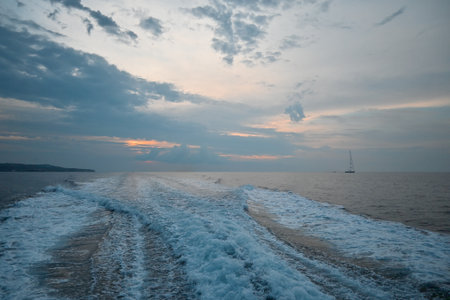 Waves trailing behind a moving boat on the sea at dusk, with dramatic clouds in the sky and a sailboat in the distance.の写真素材