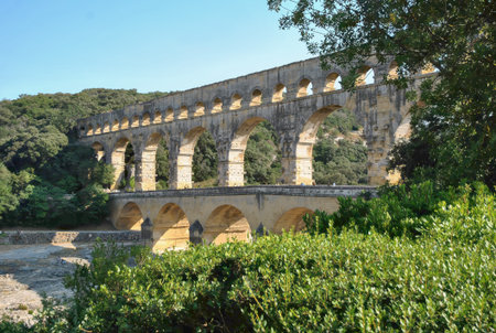Ancient Roman aqueduct bridge crossing a river, surrounded by green forest under a clear blue sky.の写真素材