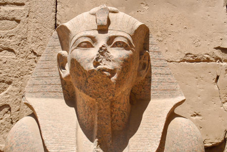 Close-up of a weathered stone statue of an ancient Egyptian pharaoh wearing a nemes headdress, with the nose and mouth partially damaged, carved into a temple wall.の写真素材