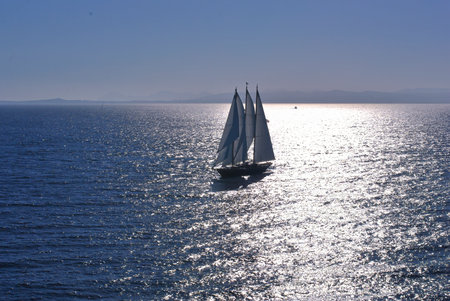 Silhouette of a sailboat on shimmering sea during late afternoon with distant coastline and clear skyの写真素材