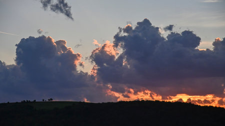 Dramatic sunset with thick clouds lit by the last rays of sunlight above a dark forested horizon.の写真素材
