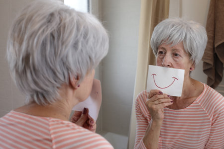 Sad senior woman holding a drawing with a smile in front of the mirror