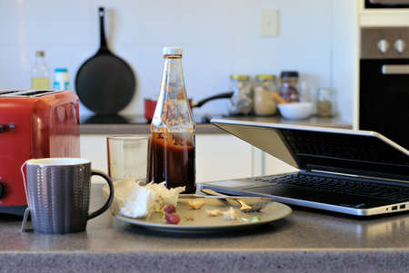 Crowded Kitchen table with laptop, plate with leftover food, tissue napkins, cup, ketchup bottle,...