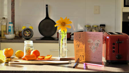 Kitchen table has a standing greeting card for mother saying 'happy mother's day' and kid's handwriting, pens, chopped vegetables and fruits on a chopping board, toaster, cooking pot, sun flower in vase, cooking pan and an alarm clock in the background.の写真素材