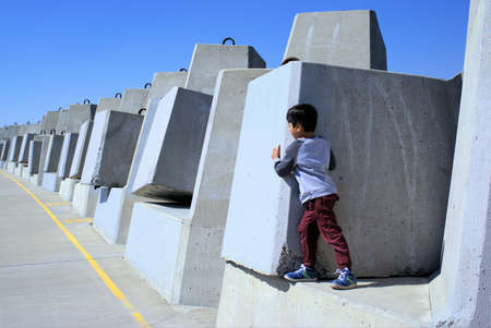 Kid peeking out from behind concrete boulder. In view is a series of concrete boulders. Boy hiding behind wall, playing hide and seek.の写真素材