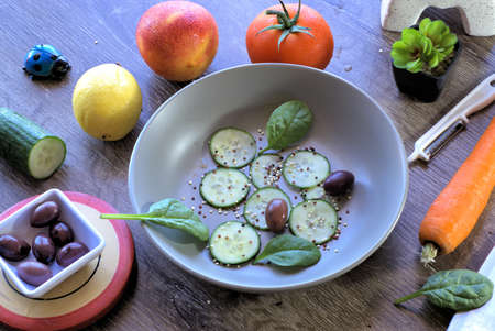 Salad preparation ingredients. Preparing fruit and vegetable salad. Bowl of cucumber slices, kalamata olives, baby spinach leaves and sprinkled quinoa. Also in image are peeled carrot, tomato, nectarine, lemon, knife, peeler, wooden chopping board and other miscellaneous objects on table.の写真素材