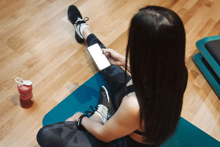 Young attractive brunette girl relaxing and holding in hands mobile phone at gym on the blue yoga...