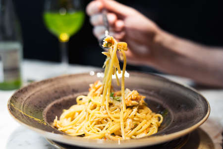 Young woman is holding fork with seafood pasta. The brown plate with seafood macaroni is in focus. Handand fork is out of focus and blurred. The glass and bottle with green basil lemonade is at the background are blurred.の写真素材