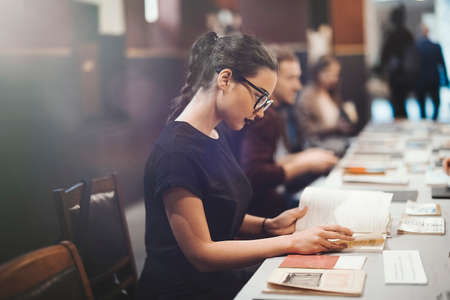 Young brunette girl with glasses and black lipstick reading book in the library. Girl is on foreground and focus, table with books is on background and blurred.の写真素材