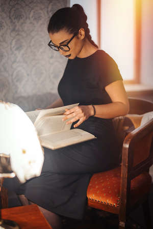 Young attractive brunette girl with glasses and black lipstick reading book on the chair.の写真素材