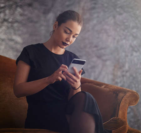 Young attractive girl holding mobile phone and texting message while sitting on the couchの写真素材