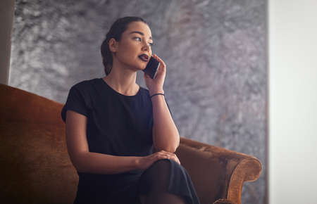 Young brunette girl talking by mobile phone while sitting on the couchの写真素材