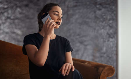 Young brunette girl talking by mobile phone while sitting on the couchの写真素材