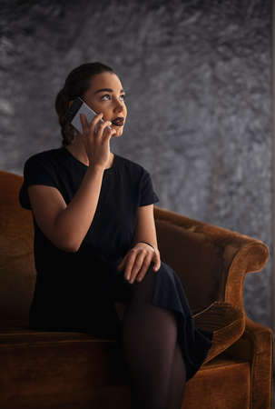 Young brunette girl talking by mobile phone while sitting on the couchの写真素材
