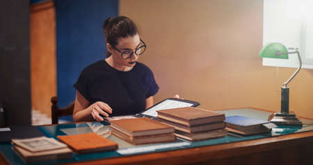 Young brunette girl with glasses and black lipstick reading book in the library. Girl is on background and focus, table with books is on foreground and blurred.の写真素材