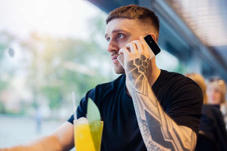 Attractive young man with tattoo is holding mobile phone and drinking fresh lemonade.の写真素材