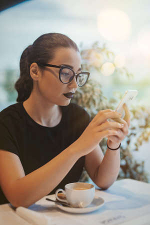 Young girl holding mobile phone it while drinking coffee in cafeの写真素材
