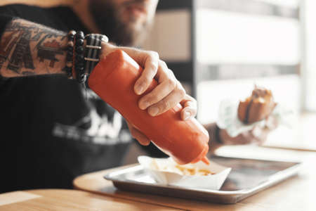 Young bearded man pours ketchup on french fries close up.の写真素材
