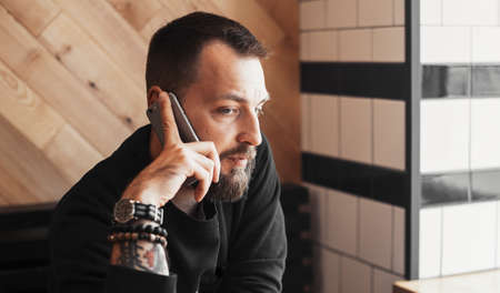 Young tattooed man talking by phone in cafe next to big window close up.の写真素材