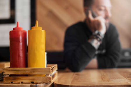 Bearded tattooed man talking by phone in cafe close up. Ketchup and mustard bottles are on foreground and focus, man is on background and blurred.の写真素材