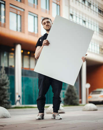 Attractive man holding blank canvas at the street and looking at the viewer.