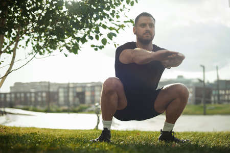 Young attractive man squatting outdoor in the park with outstretched hands.