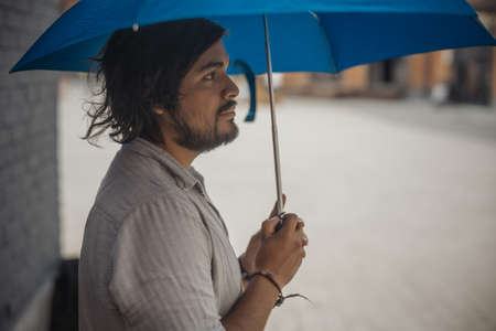 Attractive brunette latin man holding blue umbrella. Side viewの写真素材
