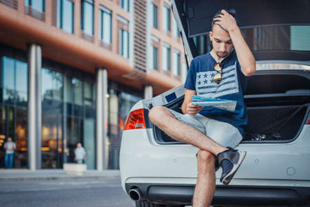 Distant plan of young man looking at map sitting in the truck of car. Man and car are on focus and foreground, background blurred. Front viewの写真素材