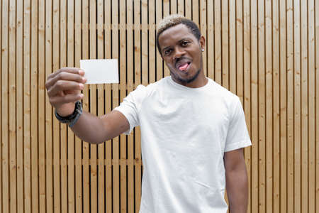 Dark-skinned man in white T-shirt showing blank business card to camera.