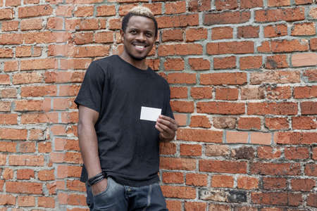 Dark-skinned man in T-shirt showing blank business card on bricked background.
