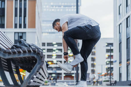 Distant plan of attractive man putting on his white sneakers and standing next to wooden bench at downtown.の写真素材