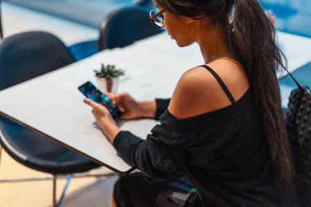 Young beautiful woman holding mobile phone in hands while waiting for her meal in modern cafe.の写真素材