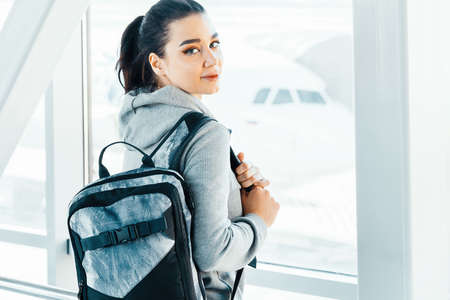 Young attractive woman with backpack standing next to big window in airport and smiling to camera.の写真素材