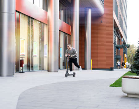 Attractive young man riding contemporary kick scooter at cityscape background.の写真素材