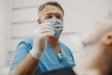 Close up of dentist in blue uniform and transparent glasses holding dental instruments and checking up teeth health condition.の写真素材