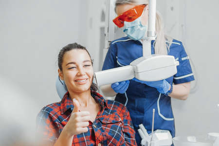Close up of woman client smiling and showing thumb up while dentist in blue uniform and orange glasses holding x-ray. Dentist is blurred, client is on focus.の写真素材