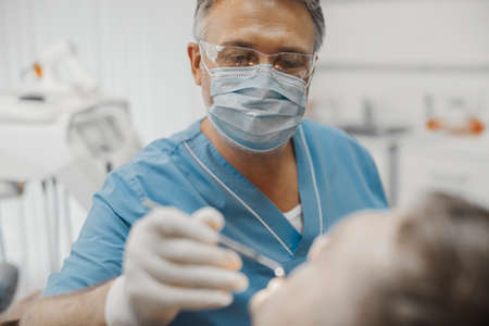 Close up of dentist in blue uniform and transparent glasses holding dental instruments and checking up teeth health condition.の写真素材