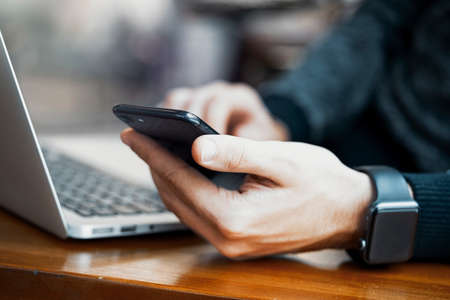 Close up of man hands with modern electronic watches and laptop drinking coffee in modern bright cafe at business lunch. Freelancer or business man working on laptop and looking at smartphone screen.の写真素材