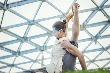 Close up of attractive athletic man practicing yoga and warming up in modern park at green grass outdoors.の写真素材