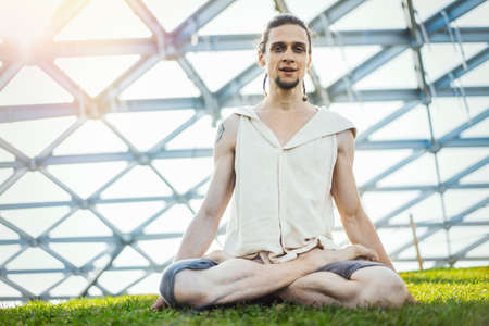 Close up of attractive athletic man practicing yoga and meditating in modern park at green grass outdoors.の写真素材