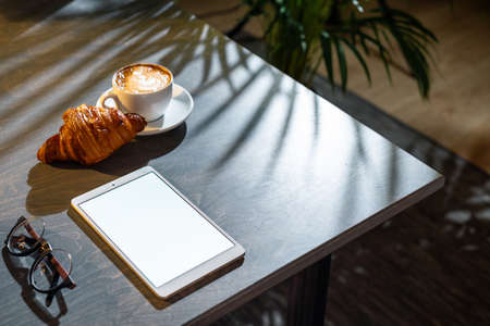 Close up of white coffee cup, croissant, glasses and pad with blank screen on dark wooden table with...