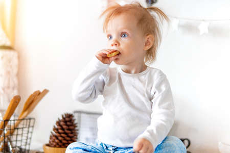 Little cute toddler girl eating homemade oat cookie and sitting on kitchen table while her elder sister preparing breakfast. Family love and care concept.の写真素材