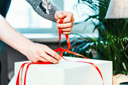 Close up of tattooed man cook ties red ribbon on white blank cake packaging. Making surprise. Hand made concept.の写真素材