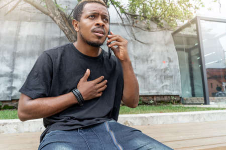 Close up of stressed dark-skinned man in black T-shirt talking by mobile phone.の写真素材