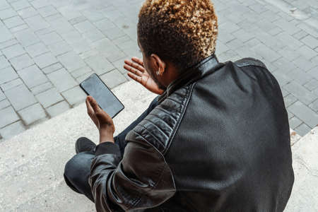 Dark-skinned man looking at mobile phone screen and sitting on concrete steps.の写真素材