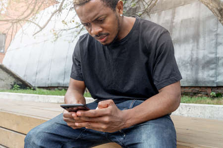 Dark-skinned man looking at mobile phone screen while sitting under treeの写真素材